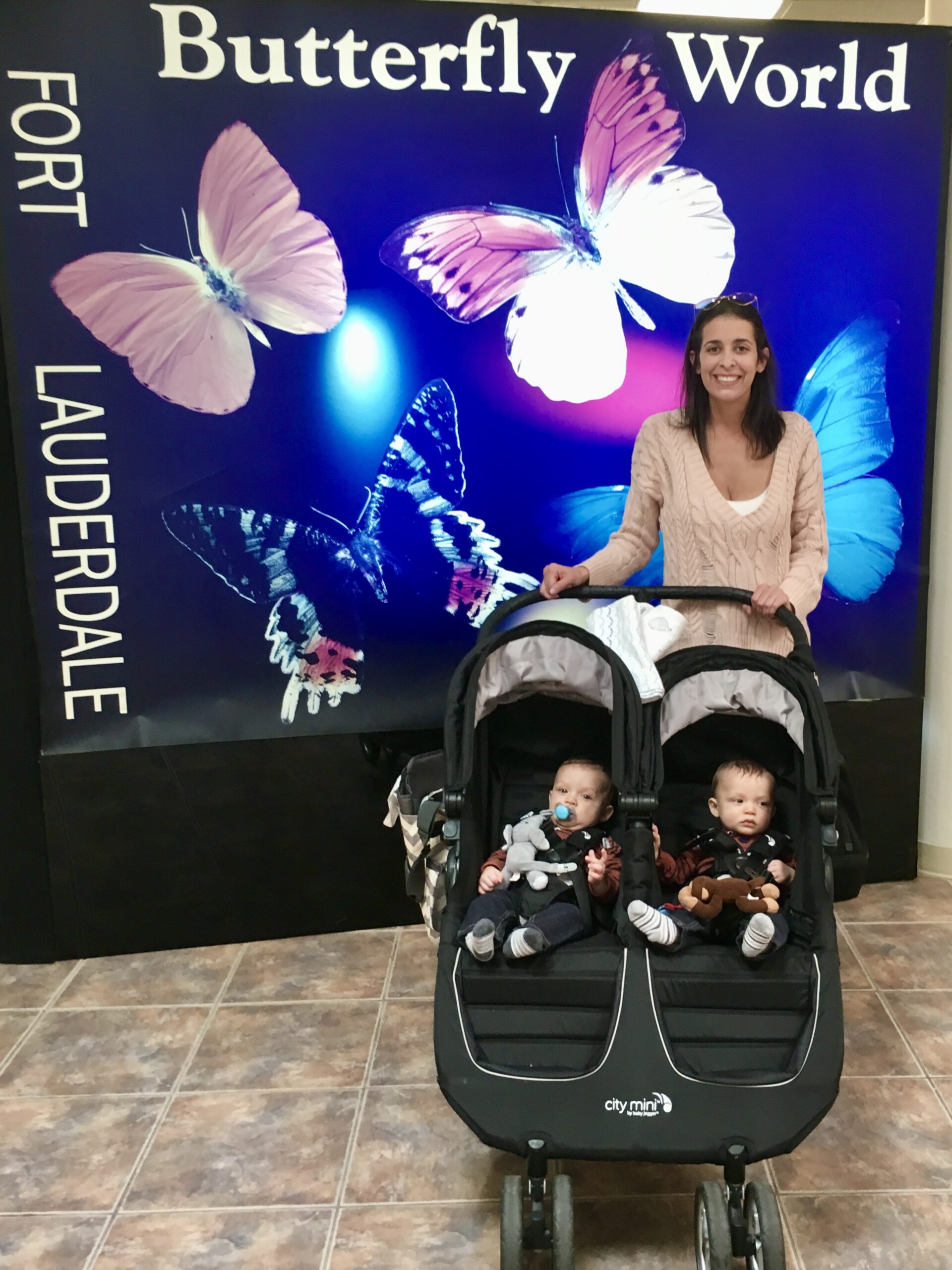 Mom posing with twin infant boys in a double stroller in front of the glowing Butterfly World entrance sign in Coconut Creek, Florida