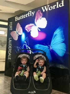 Twin toddler boys in a double stroller smiling in front of the Butterfly World Fort Lauderdale entrance sign